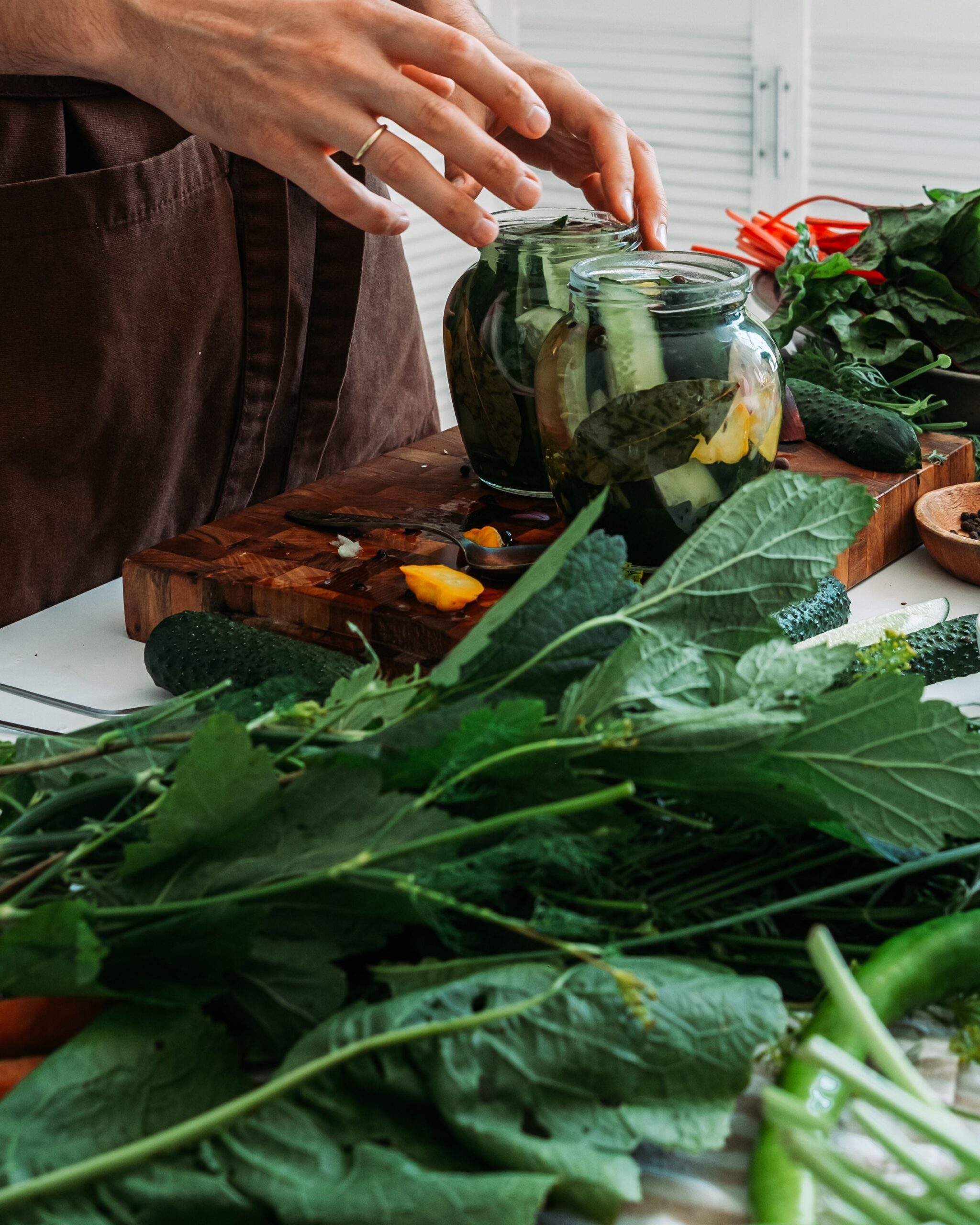 Glass pickling jars with cucumbers and fresh garden herbs on a wooden cutting board