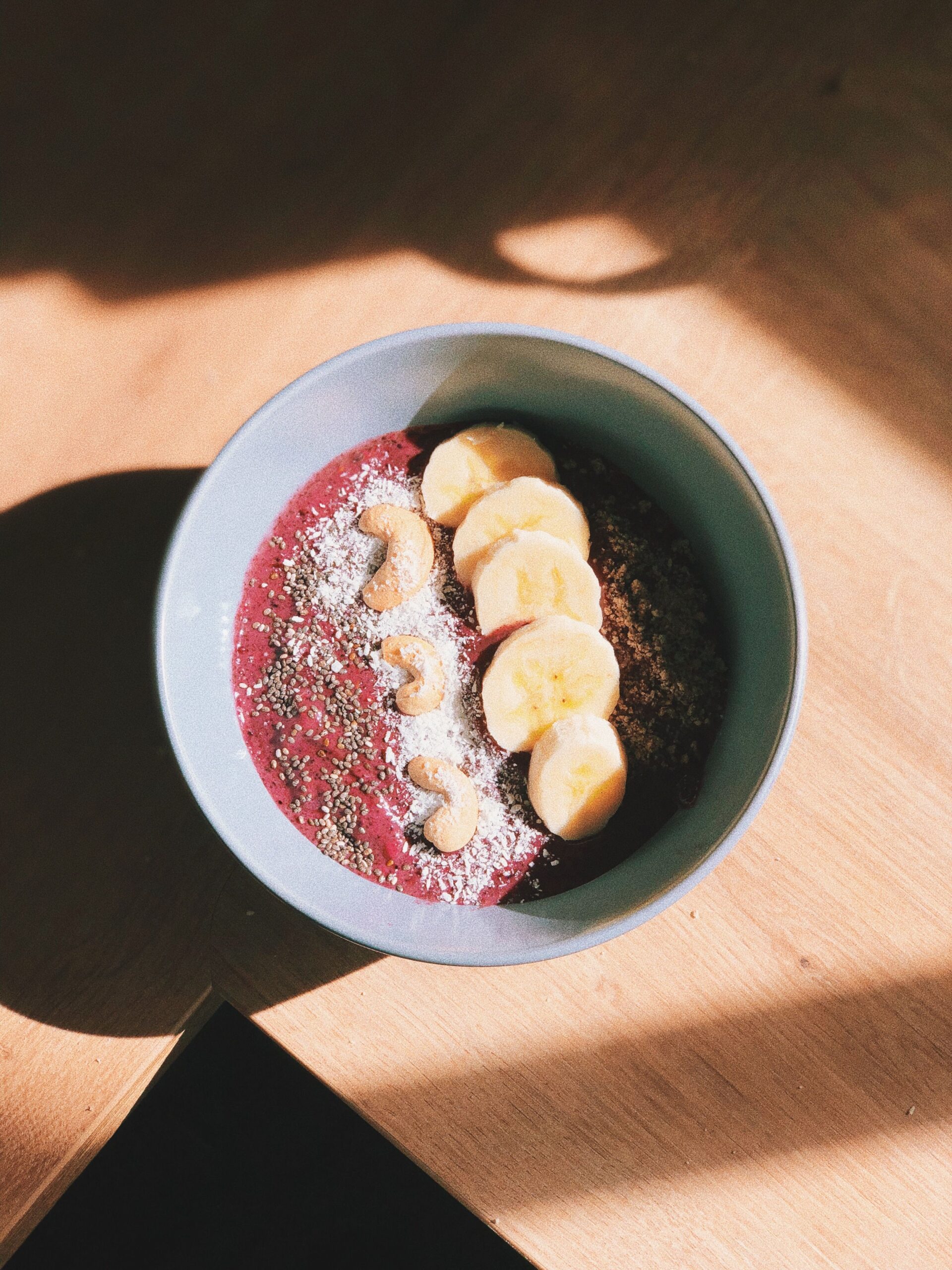 Overhead shot of a berry smoothie bowl decorated with sliced banana, cashews, shredded coconut, and chia seeds